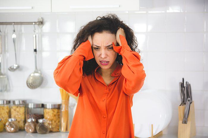 Woman in an orange shirt looking frustrated in a kitchen, upset about someone disliking her favorite pizza place.