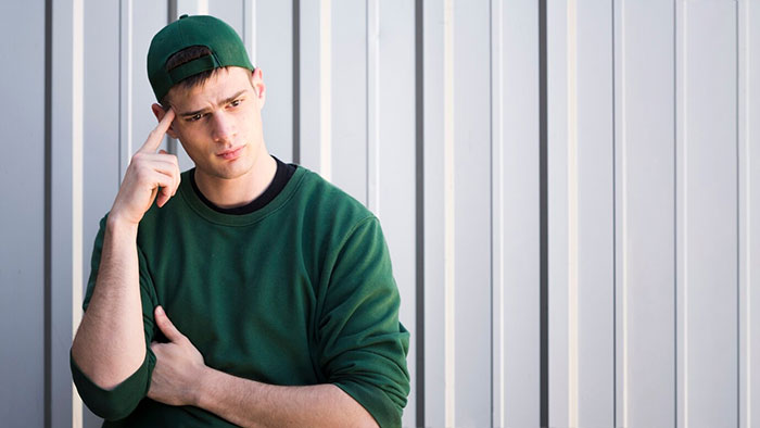 Teen brother in green sweater and cap, looking thoughtful against a metal wall.