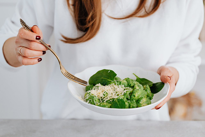 Woman in a white sweater holding a plate of green gnocchi with a fork, garnished with cheese and spinach.