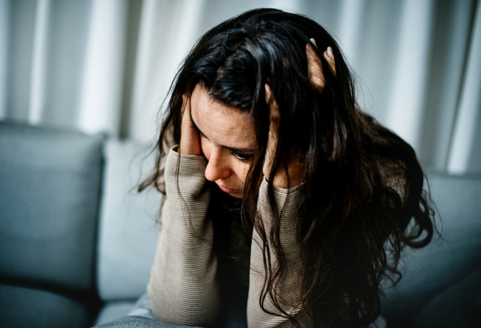 A distressed woman with long brown hair sitting on a couch, holding her head in her hands.