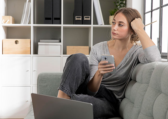 Woman pondering Christmas party attendance, sitting on a couch with a phone, concerned about gift price.