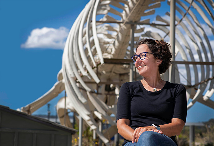 Colossal Biosciences Chief Science Officer Beth Shapiro smiling in front of large dinosaur skeleton under a clear blue sky.