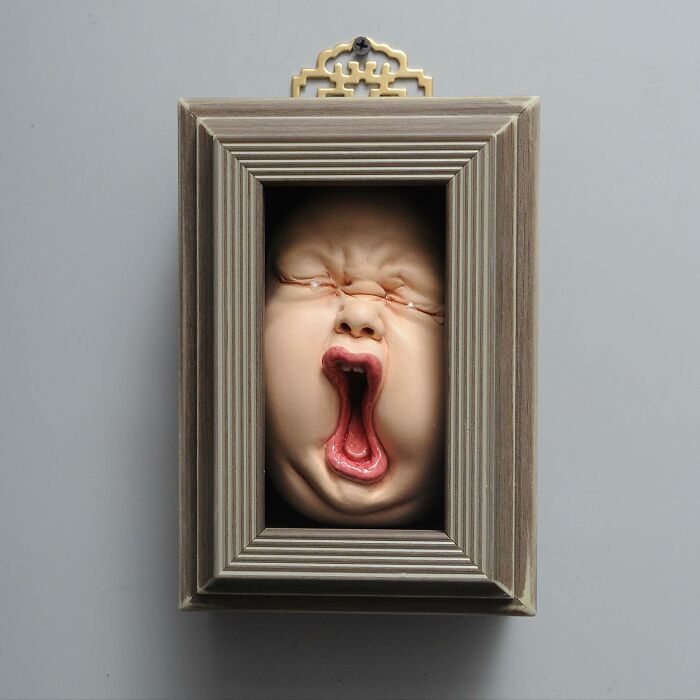 A framed sculpture blending realism and surrealism, featuring an expressive human face with an open mouth.