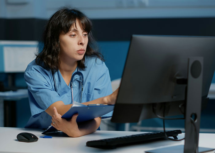 Nurse working on a computer, looking focused, while holding a clipboard in a hospital setting.