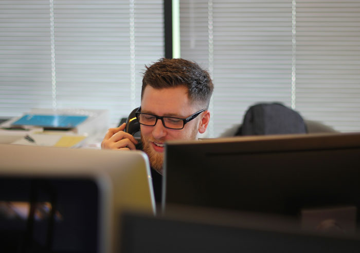 Man in an office environment, smiling while on a phone call, with computer screens in front of him.