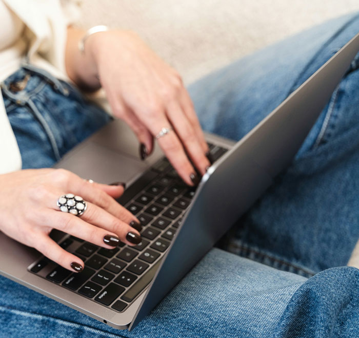 Gen A individual using a laptop, wearing a decorative ring, and jeans, engaged in technology interaction.