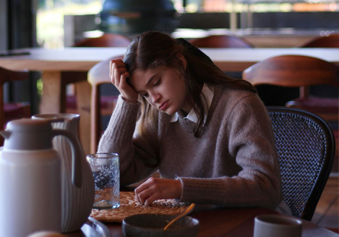 A Gen A girl looking frustrated at a table with glassware, resting her head on her hand in a cozy setting.