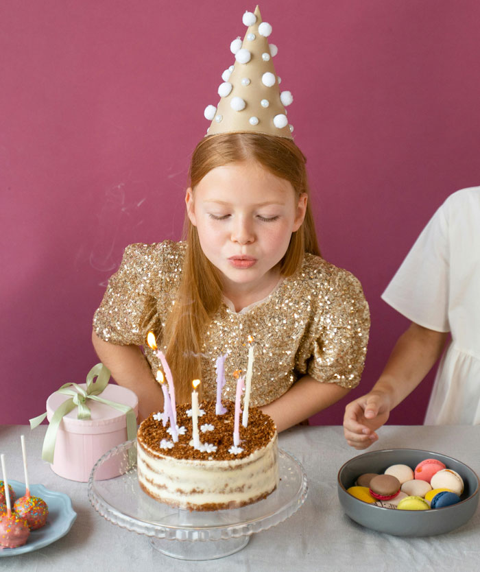 Girl in a party hat blowing out candles on a cake during a birthday celebration.