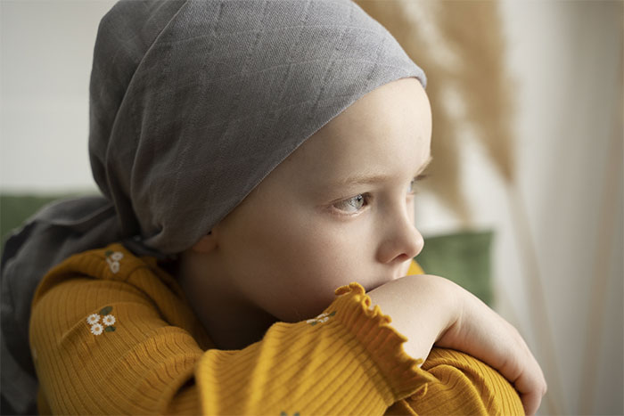 Teen girl with a headscarf, wearing a mustard sweater, gazing out a window thoughtfully.