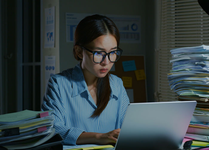 Woman using a laptop in an office, surrounded by stacks of documents, working late as focus shifts to technology use by Gen A.