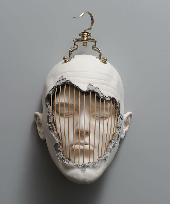 Sculpture blending realism and surrealism shows a face with a broken facade and metal cage-like structure, hung by a hook.