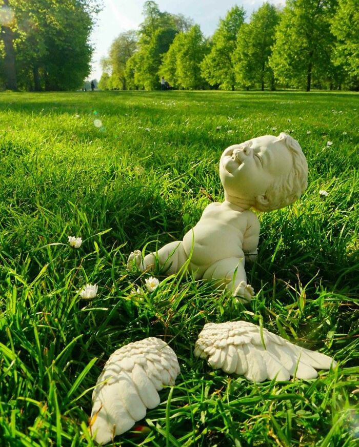 Sculpture blending realism and surrealism of a baby with wings on grass in a park.