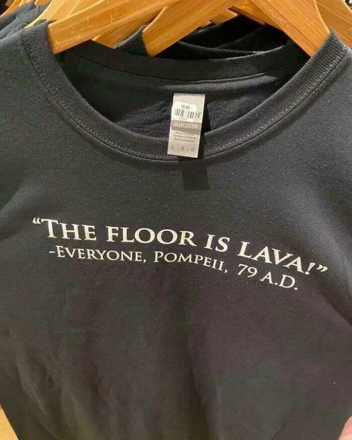 Black t-shirt with funny text "The floor is lava! - Everyone, Pompeii, 79 A.D." displayed on a hanger.