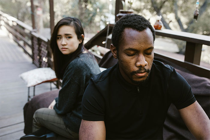 Man refusing to take back cheating girlfriend, sitting apart on an outdoor bench with a distant expression.