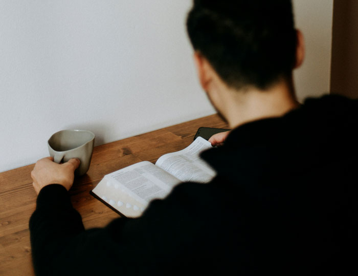 Man reading a book at a wooden table, holding a coffee mug, focusing on finals, not babysitting sister's kids during vacation.