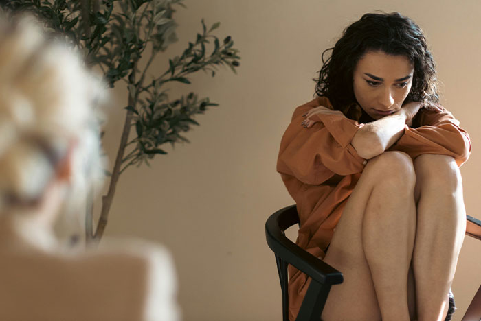 Woman sitting on a chair, appearing thoughtful and concerned, amidst a conversation about financial and family issues.