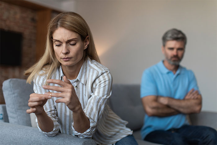 A woman looks at her ring, deep in thought, while a man in a blue shirt sits in the background, arms crossed, highlighting ex-husband regrets.