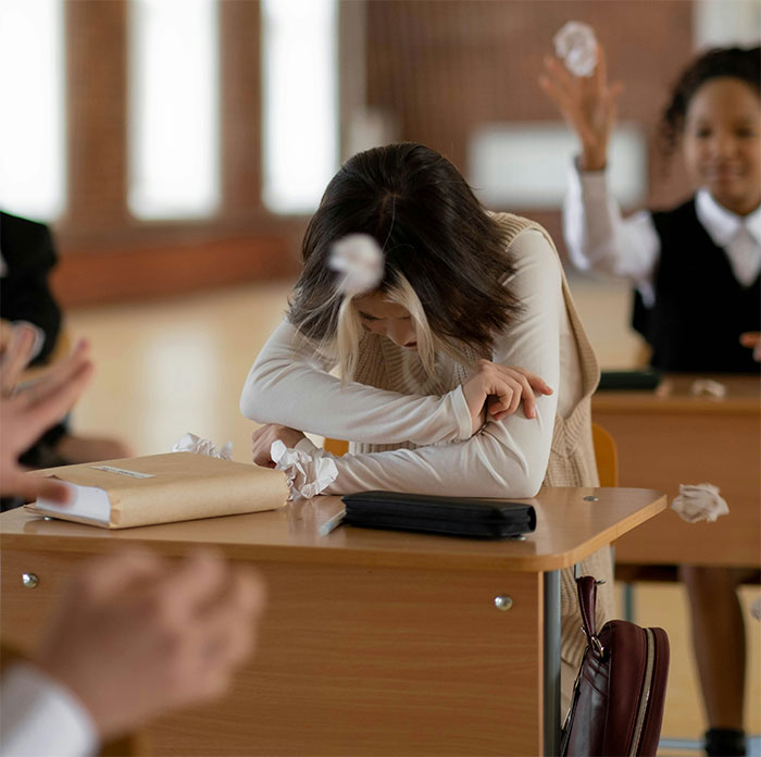 Teen looking uncomfortable in a classroom setting, covering head while being targeted with paper balls.