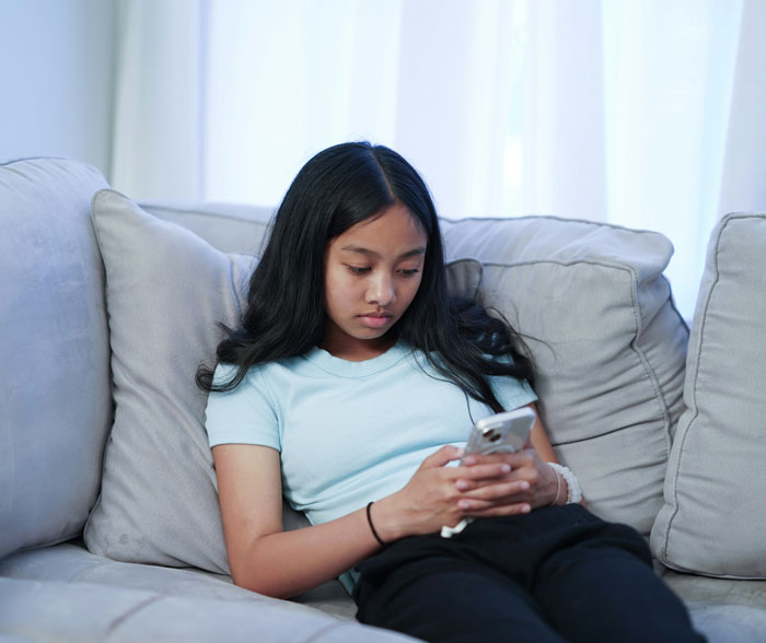 Gen A girl using a smartphone while sitting on a couch, wearing a light blue shirt, and focused on the screen.