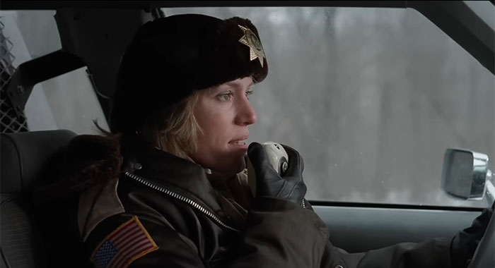 Strong female protagonist in uniform driving, using a radio in a snowy setting.