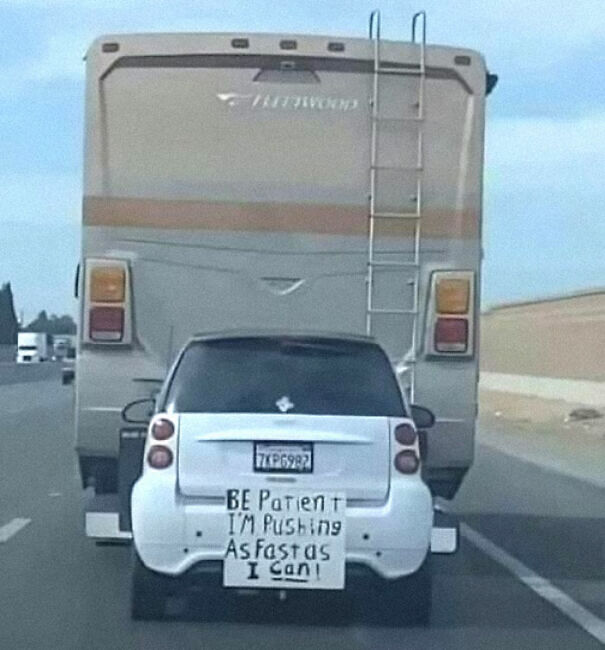 Absurd sign on car rear says, "Be patient I'm pushing as fast as I can," behind large RV on highway.