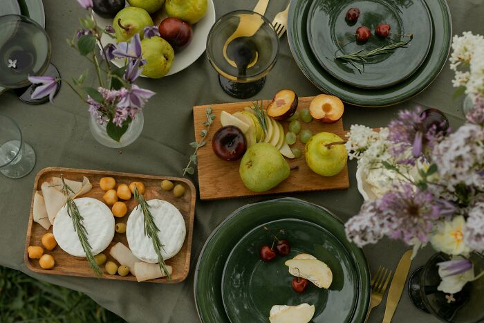 Frugal home hack: elegant picnic setup with green plates, fresh fruits, cheese, flowers, and gold utensils on a green cloth.