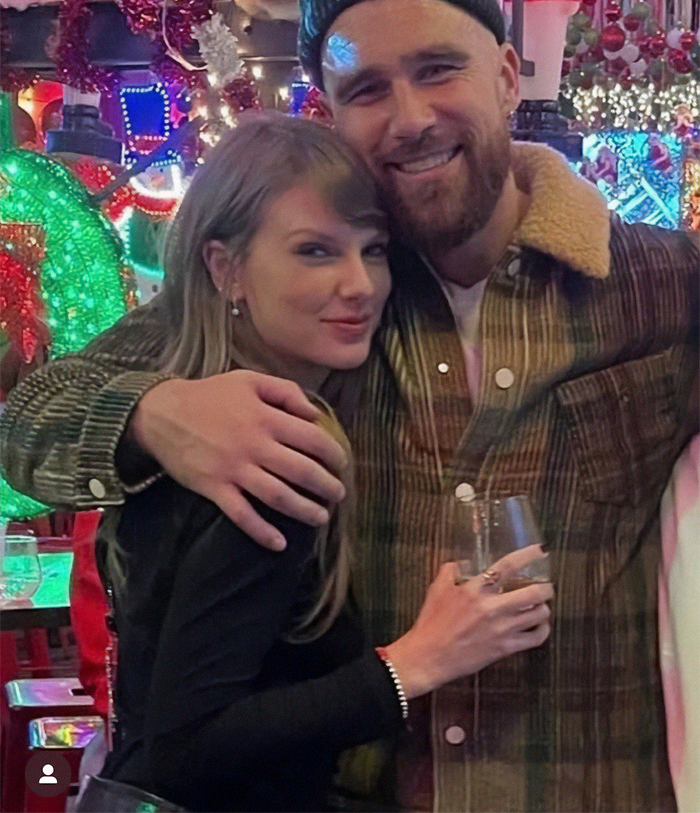 Taylor Swift smiling at an event, wearing a black outfit, amid festive decor, with Travis Kelce in a plaid jacket.