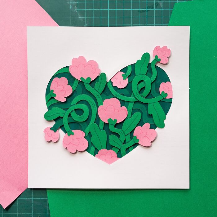 Layered paper illustration of a heart with pink flowers and green leaves on a crafting table.