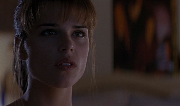 Strong female protagonist looking determined, dimly lit room, intense expression, with artwork blurred in the background.