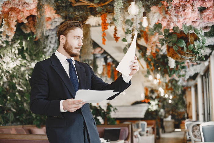 Man in suit at a wedding venue, looking at papers, surrounded by floral decorations.