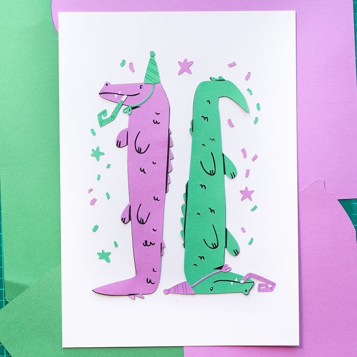 Layered paper art illustration of cute crocodiles in party hats, with confetti, on a white background surrounded by green and purple paper.