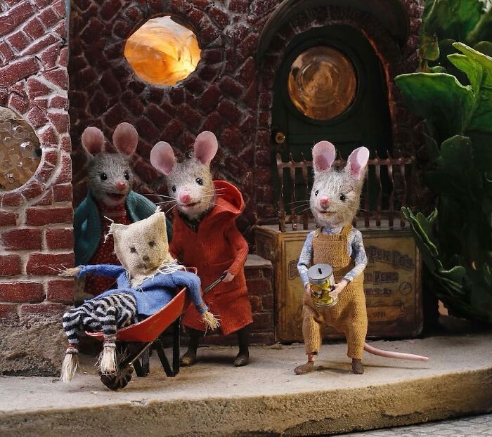 Miniature mice in colorful outfits with a wheelbarrow in front of a charming brick building from Mouseland.