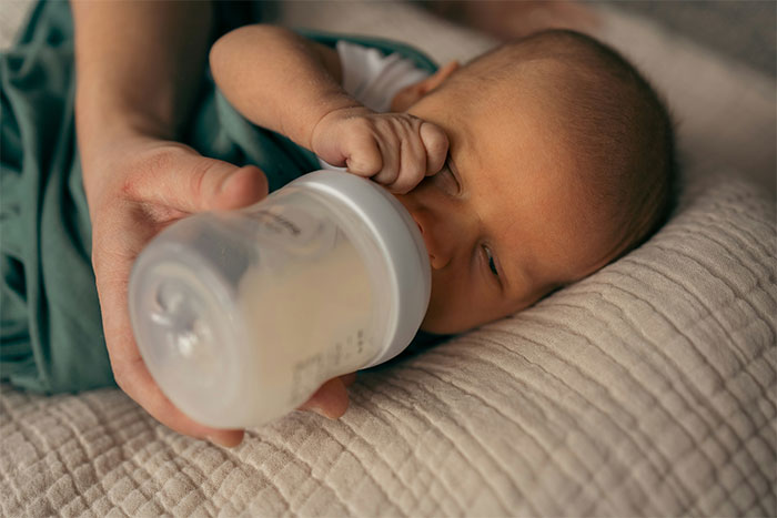 A baby being bottle-fed while lying on a blanket, related to a woman's decision about her mother-in-law seeing the baby.