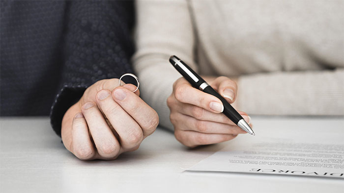 Hands holding a wedding ring and a pen, contemplating divorce papers.