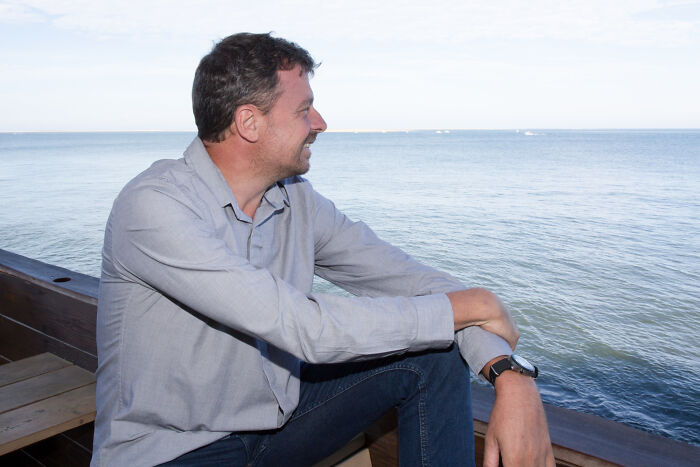 Man in a casual shirt sitting by the ocean, deep in thought.