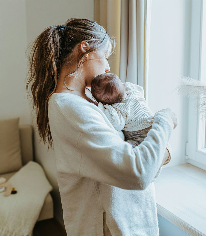 Woman holding baby by a window, wearing a light sweater, showcasing a moment of tenderness and care.