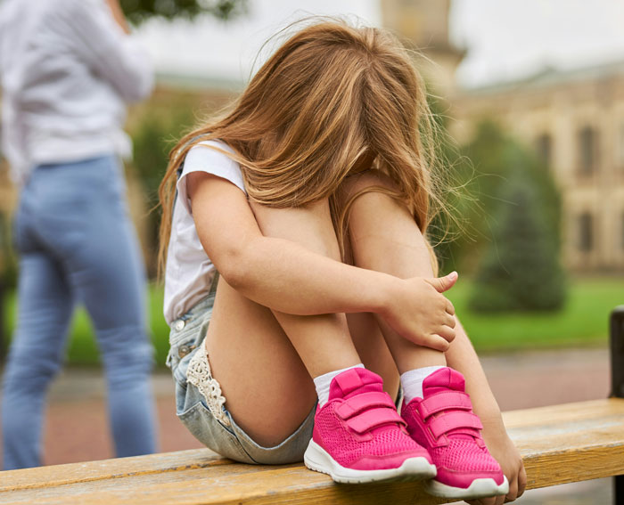 Young Gen A girl with long hair seated on a bench, wearing pink sneakers, appearing thoughtful outdoors.