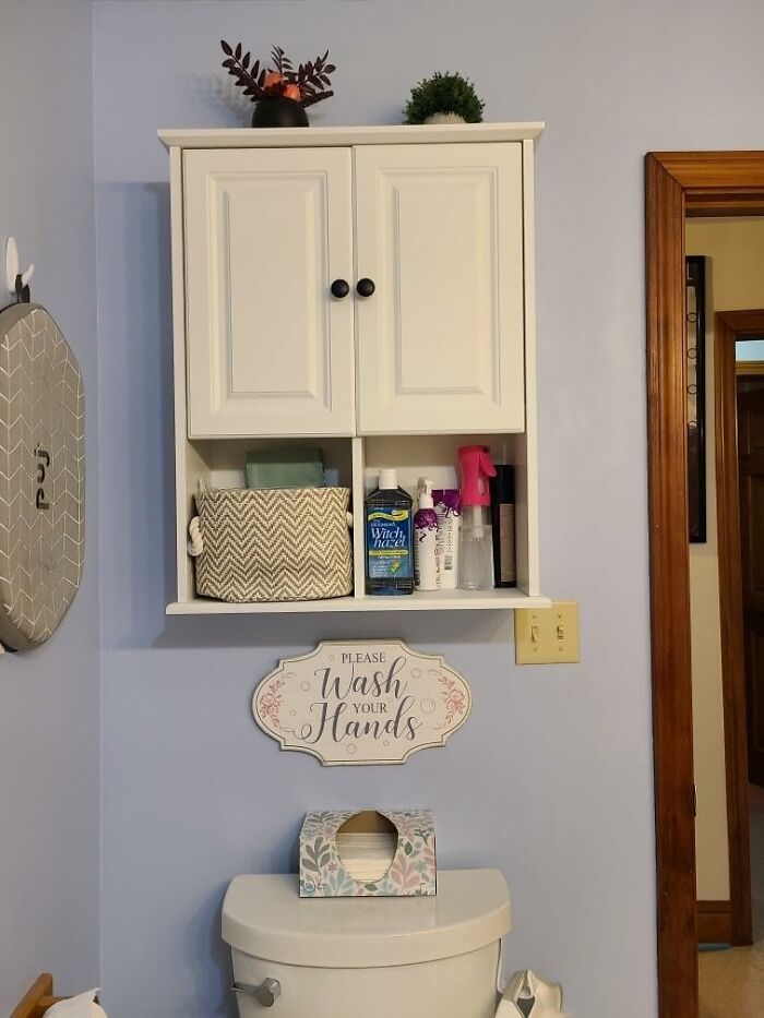 White bathroom cabinet with plants and cleaning supplies, above a toilet with a "Wash Your Hands" sign.