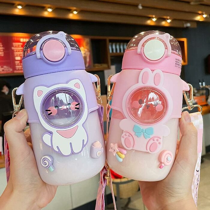 Cute cartoon-themed water bottles, one purple with a cat design and one pink with a bunny design, held up in a store.