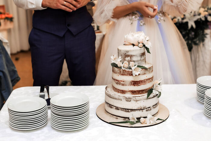 Wedding couple standing behind a floral cake, with plates stacked nearby on a table.