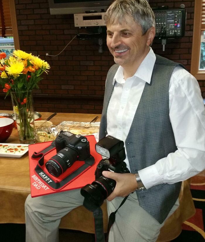 Man holding cameras at a party, smiling next to a table with flowers, embodying hilarious in-laws humor.