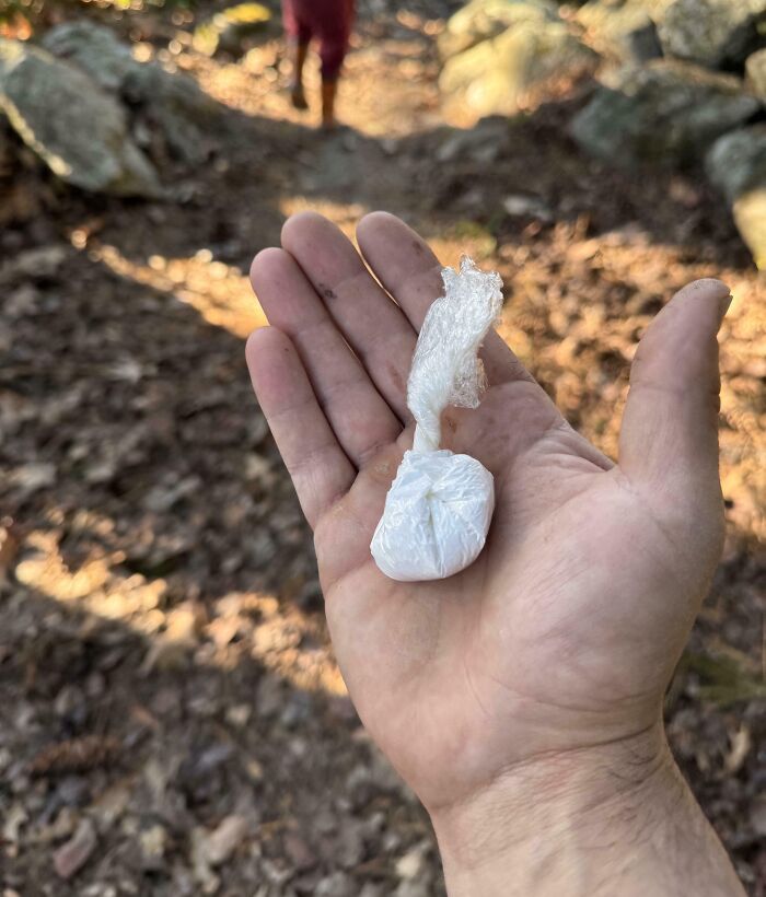 A hand holding a small bag wrapped in plastic, outdoors on a dirt path. Hilarious-In-Laws.