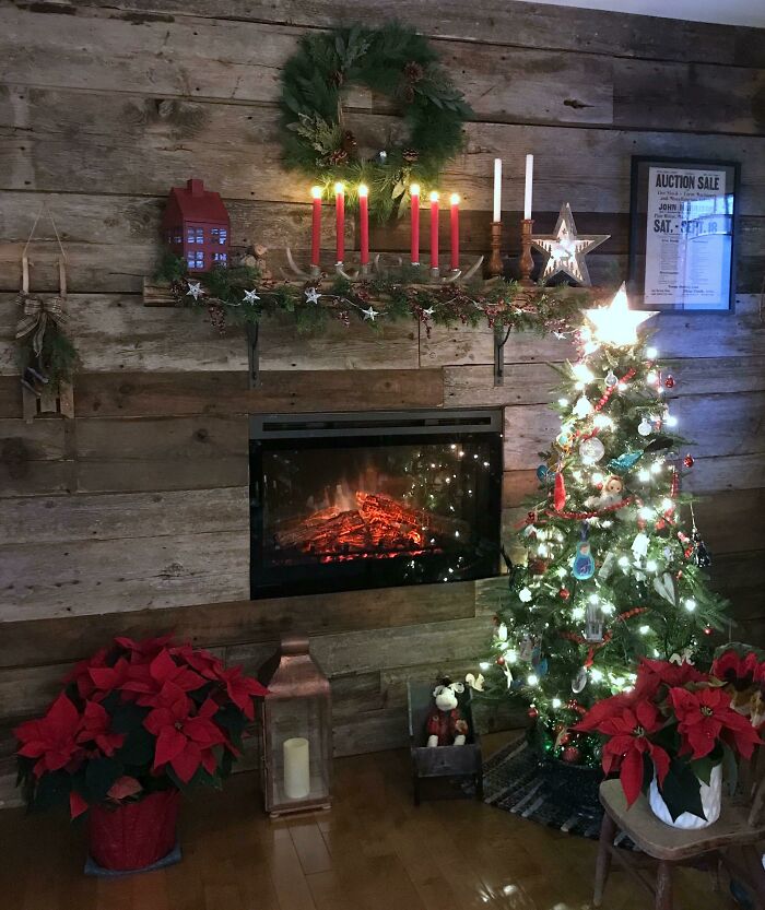 Rustic fireplace with Christmas decorating ideas featuring a lit tree, poinsettias, and festive wreath on a wooden wall.