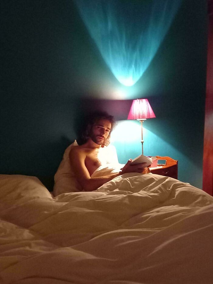 Man in bed under soft lamp light, holding a book, resembling Renaissance painting ambiance.