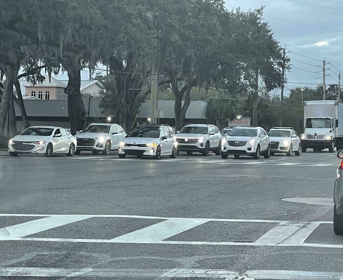 Cars lined up at a traffic light on the road, with trees in the background and a cloudy sky above.