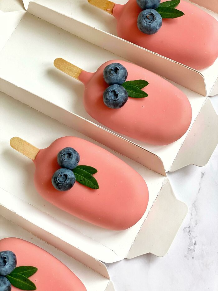 Pink desserts with blueberries and green leaves on top, arranged neatly in white packaging. Beautiful food presentation.