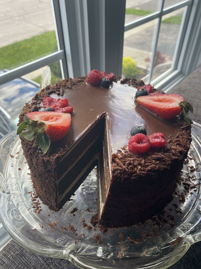 Chocolate cake with berries on top, featuring sliced strawberries, raspberries, and blueberries, on a glass plate.