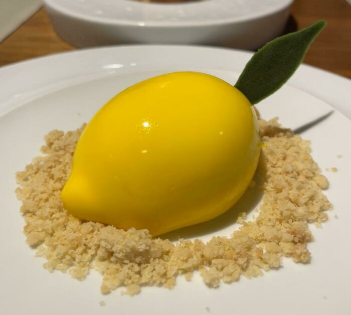 A lemon-shaped dessert with a glossy yellow finish, garnished with crumbs on a white plate, perfect for foodies to love.