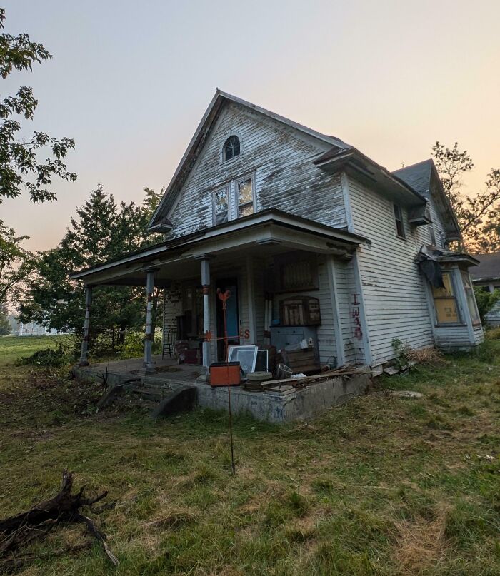 Old inherited house with a weathered exterior, surrounded by trees and overgrown grass at sunset.