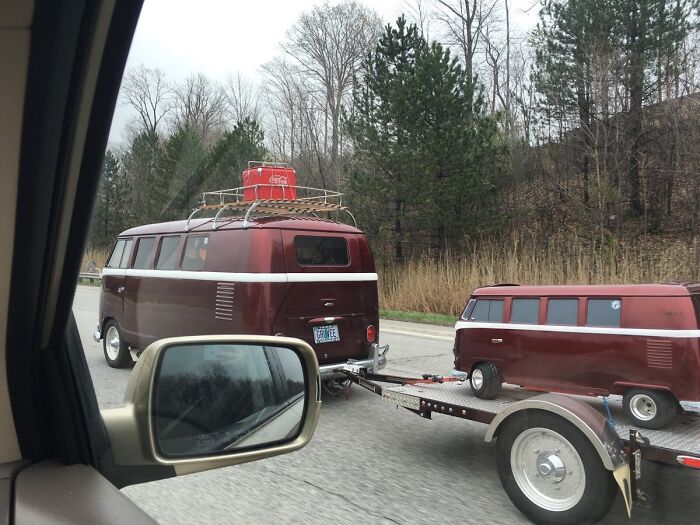 Red vintage van towing a miniature version on a trailer, spotted on the road.